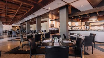 A spacious restaurant with high ceilings, wooden beams, and a central kitchen area. Tables are set with white tablecloths and black chairs.