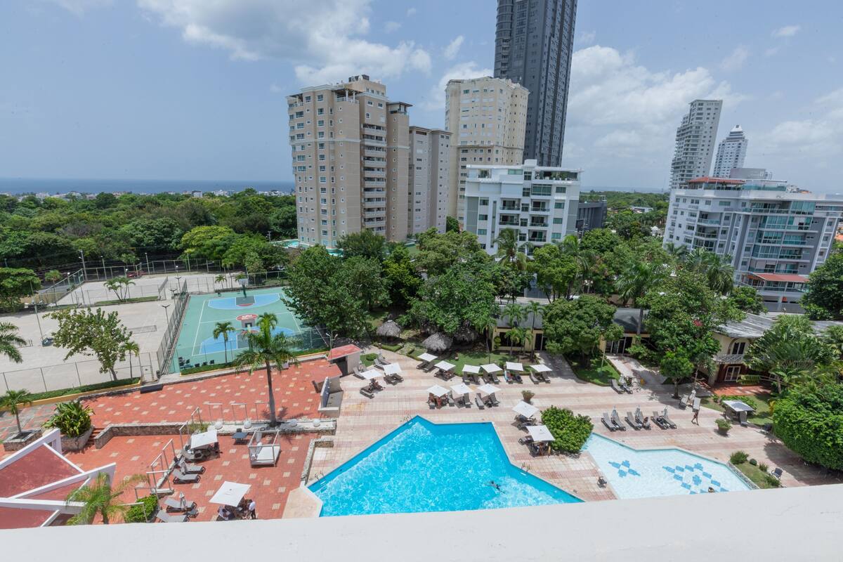 A rooftop pool area with multiple swimming pools, lounge chairs, and a view of surrounding high-rise buildings.
