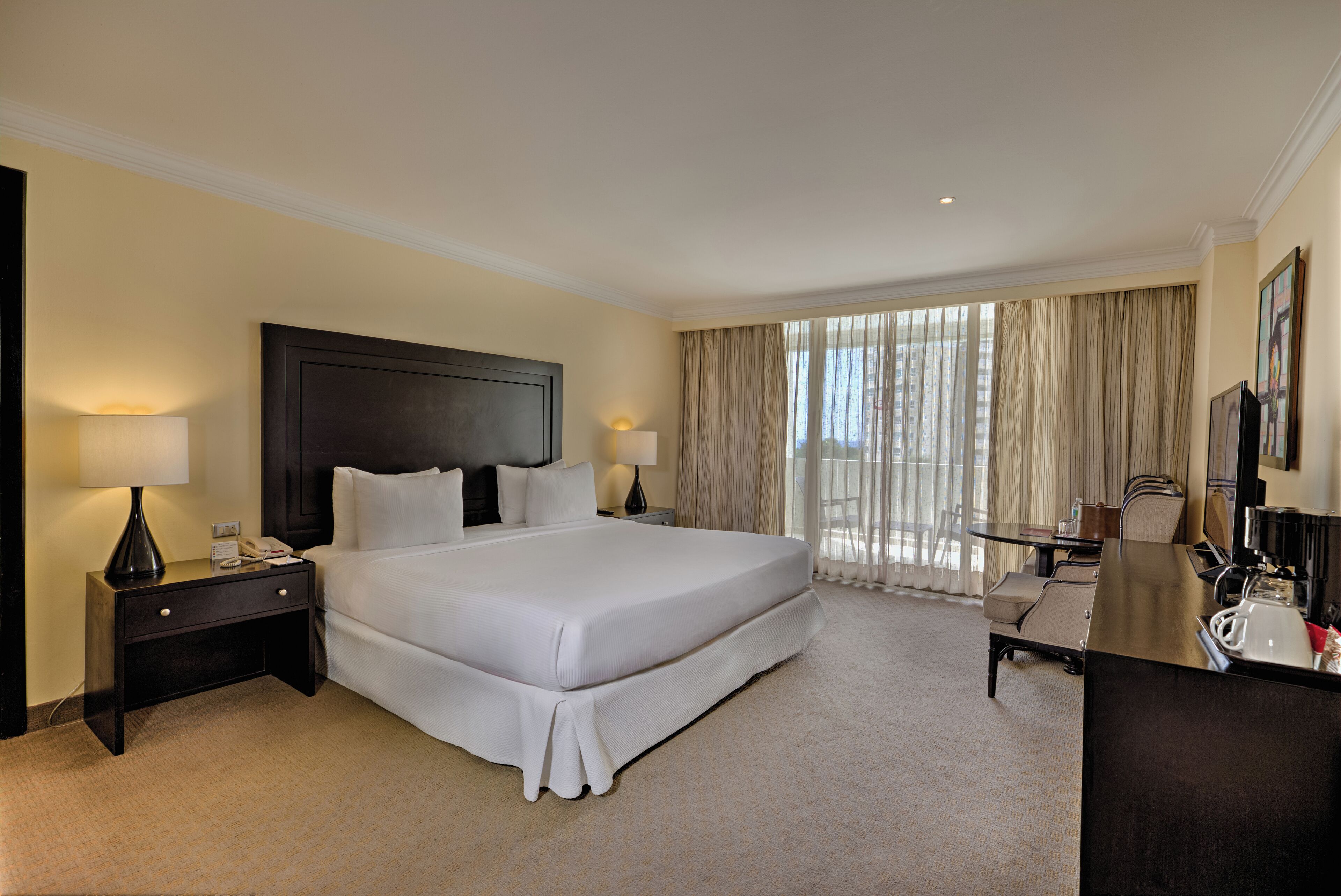 A spacious hotel room with a large bed, two bedside tables with lamps, a desk with a chair, and a large window with curtains.