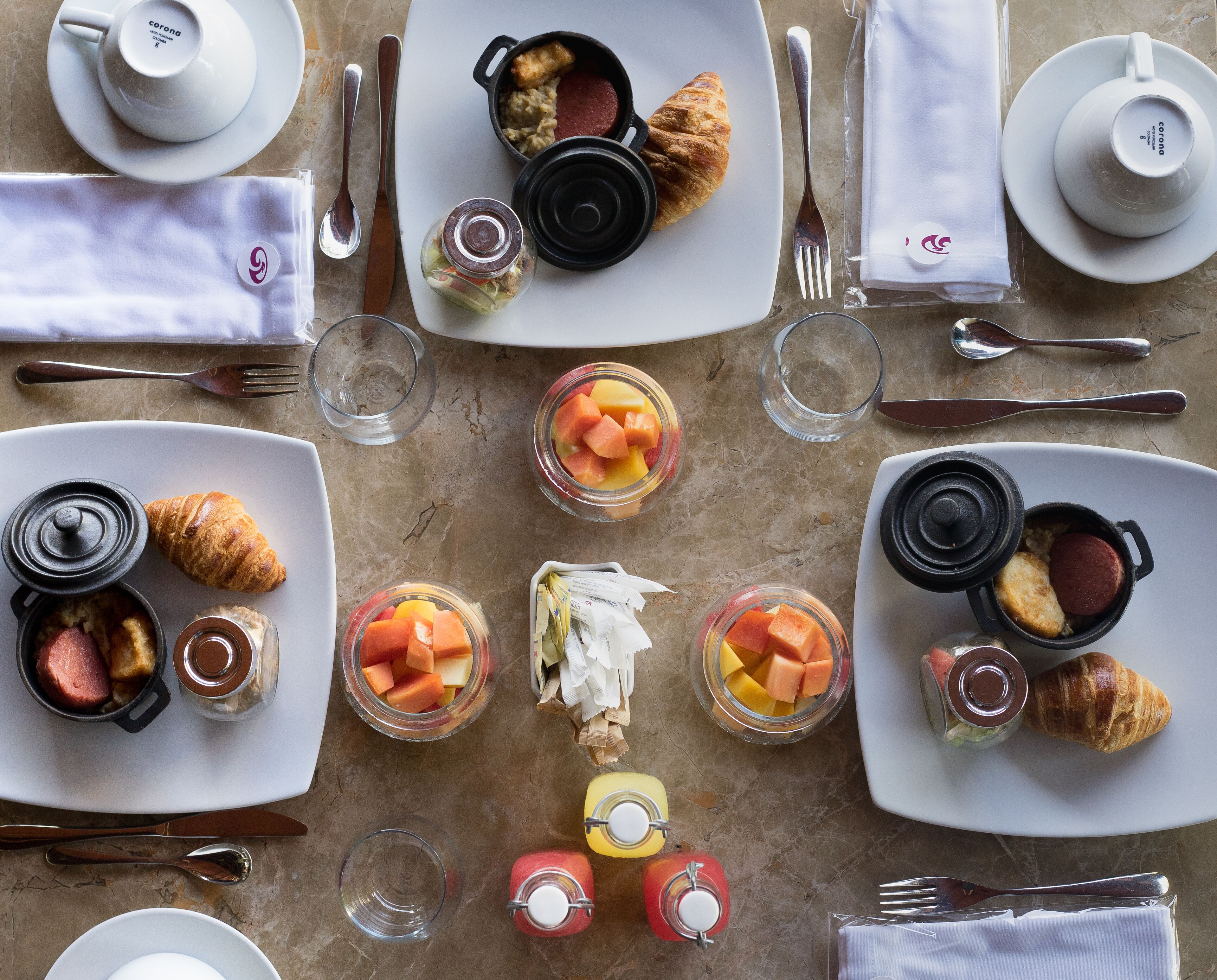 A breakfast setting with croissants, fruit, and condiments on plates, utensils, and napkins.