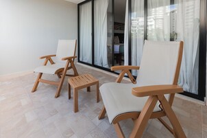 Un balcón con tres sillas de madera, una mesita pequeña, puertas de cristal amplias y vistas a edificios.