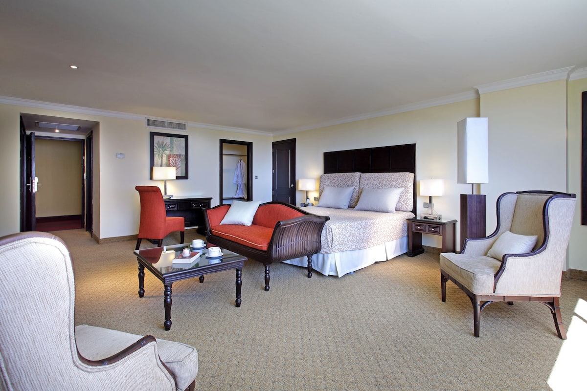 A hotel room with a bed, two chairs, a coffee table, a desk, and a lamp.