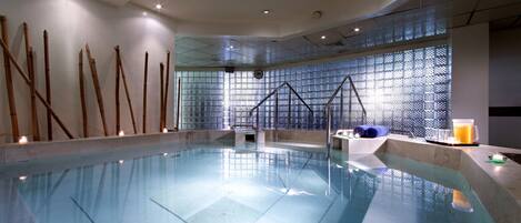An indoor swimming pool with clear water, a sunlit area with a window, and a seating area with a drink.