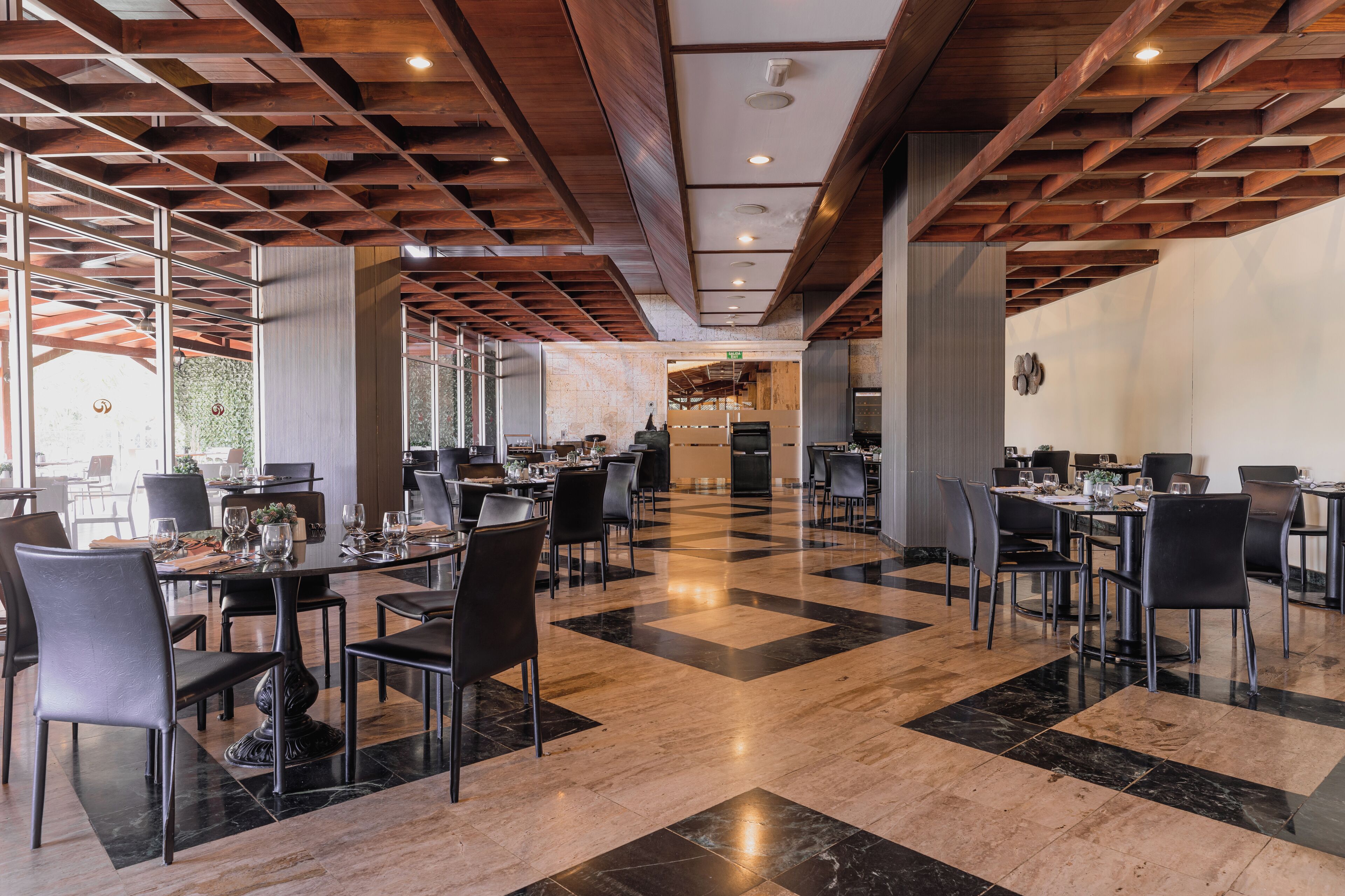 A spacious restaurant with high ceilings, wooden beams, and a checkered floor pattern. Tables are set with chairs, and the space is well-lit.