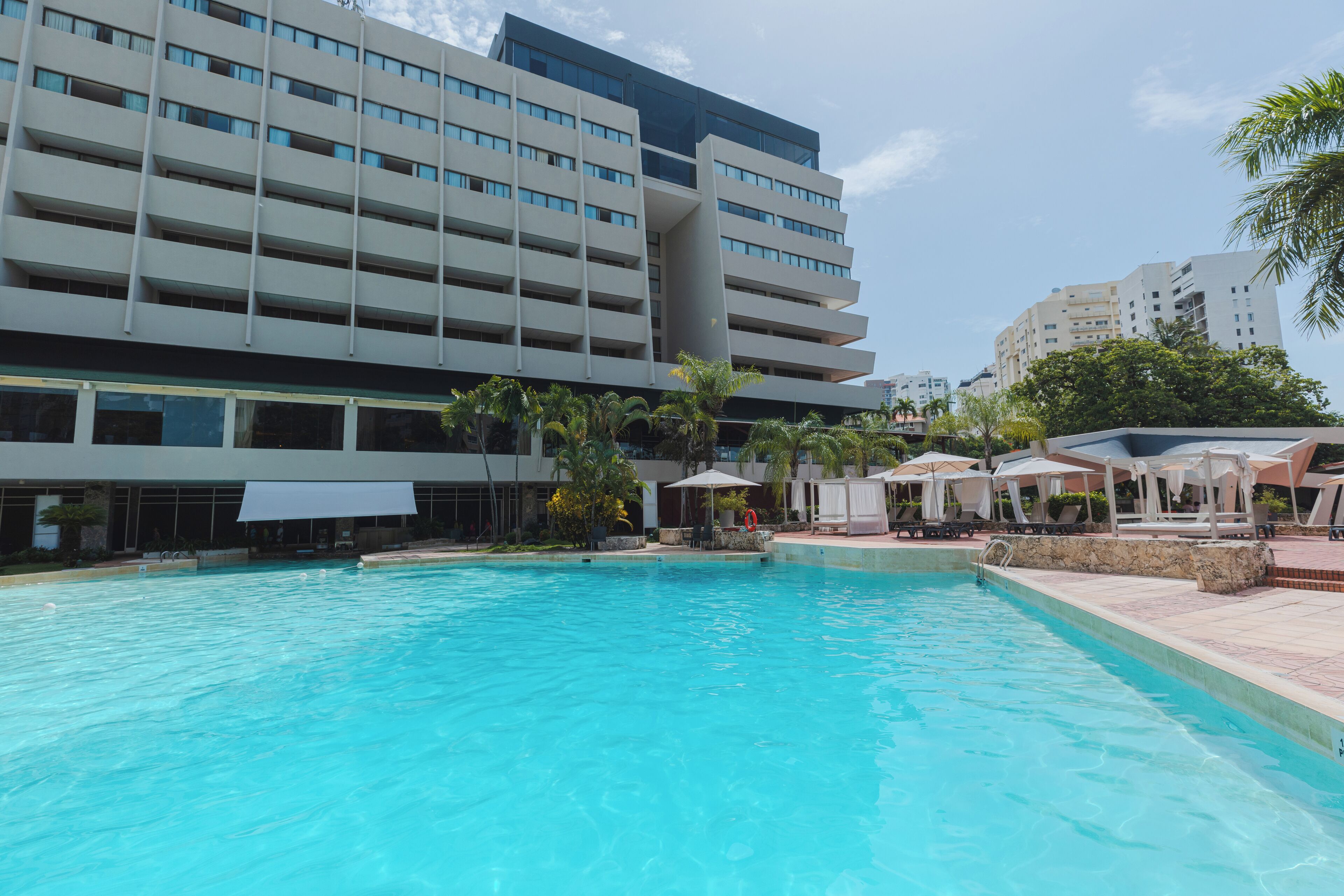 A swimming pool with lounge chairs and umbrellas, surrounded by a modern building with multiple floors and balconies.