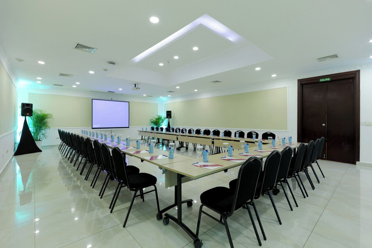 Un salón de conferencias con una larga mesa, sillas, una pantalla de proyector y una puerta con la etiqueta 'SALIDA'.