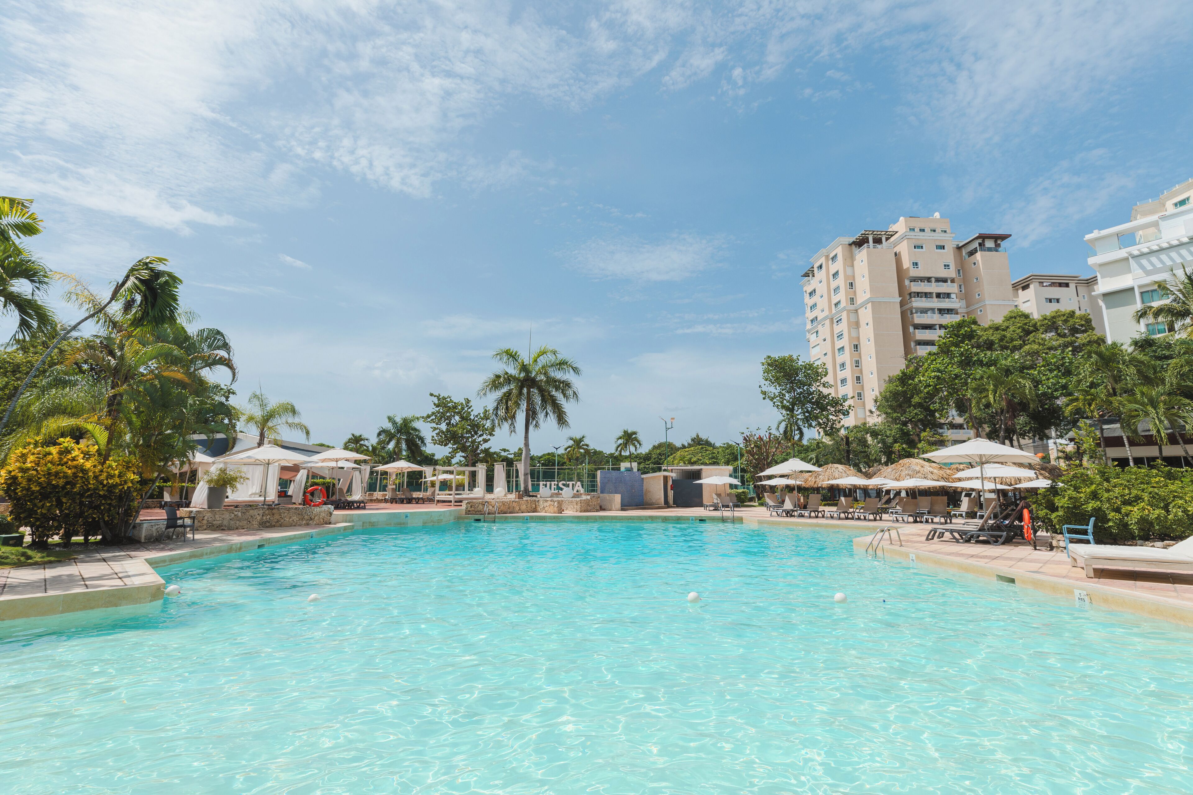 A swimming pool with lounge chairs and umbrellas, surrounded by palm trees and buildings.