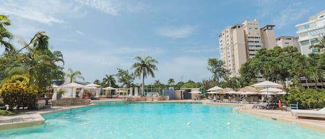 A swimming pool with lounge chairs and umbrellas, surrounded by palm trees and buildings.