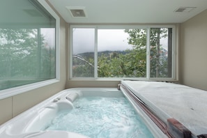 A room with a hot tub, large windows, and a view of trees and hills.