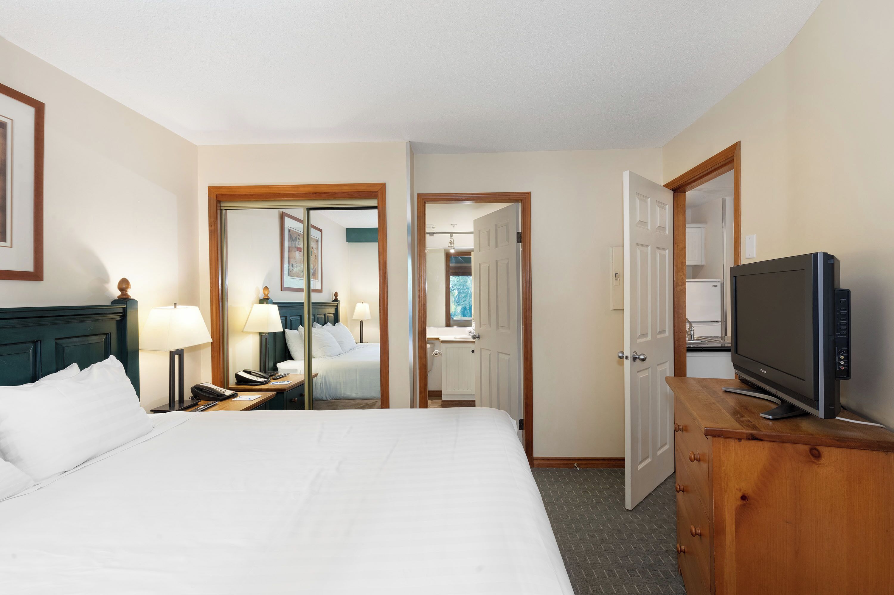 A hotel room with a bed, bedside lamps, a TV on a wooden dresser, and a bathroom visible through an open door.