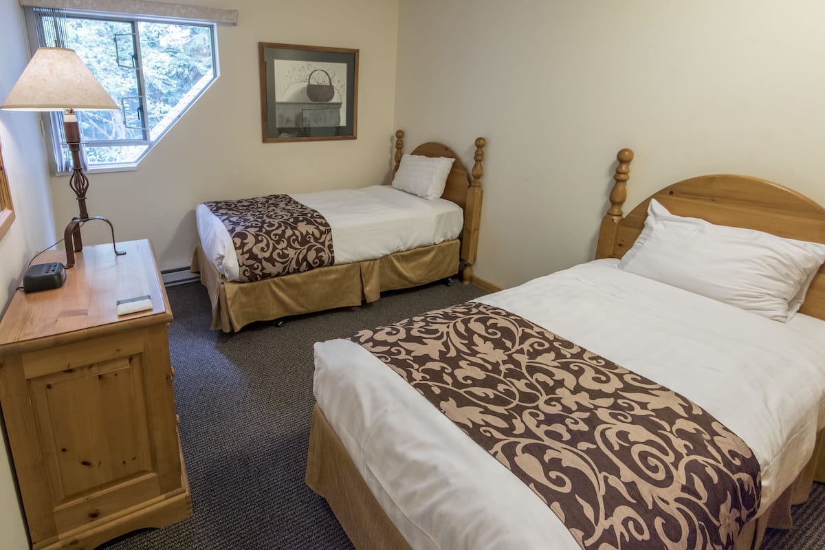 A hotel room with two beds, a nightstand, a lamp, and a window with a view of trees.