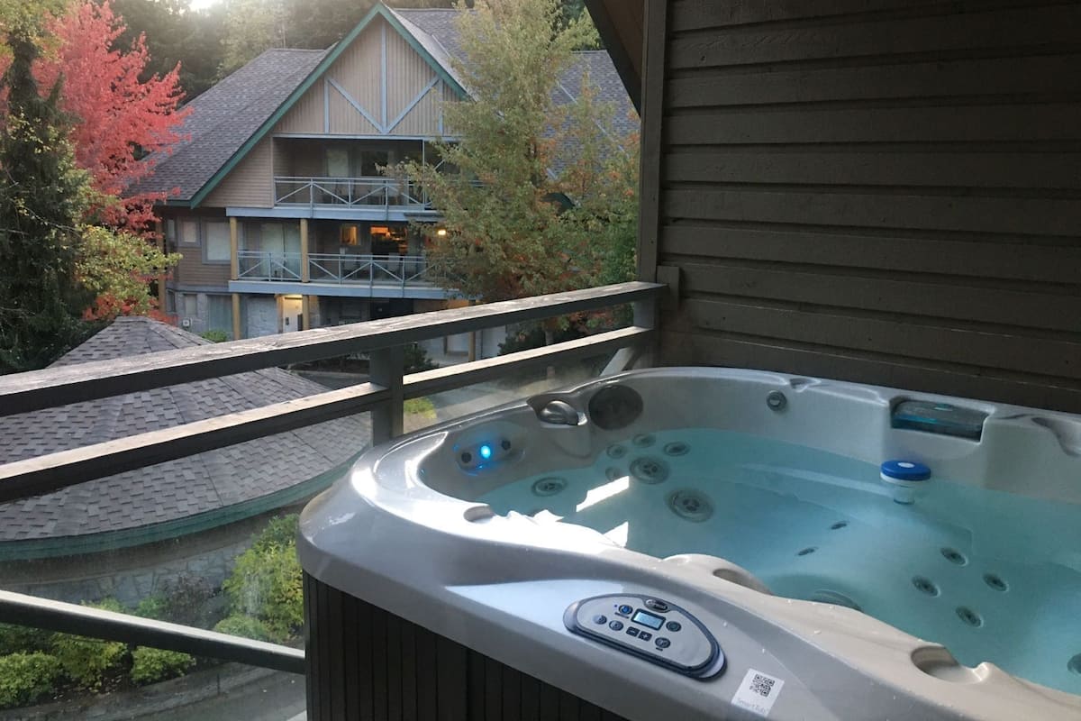 A hot tub with a control panel, a balcony railing, and a view of a building with a blue railing and a sloped roof.