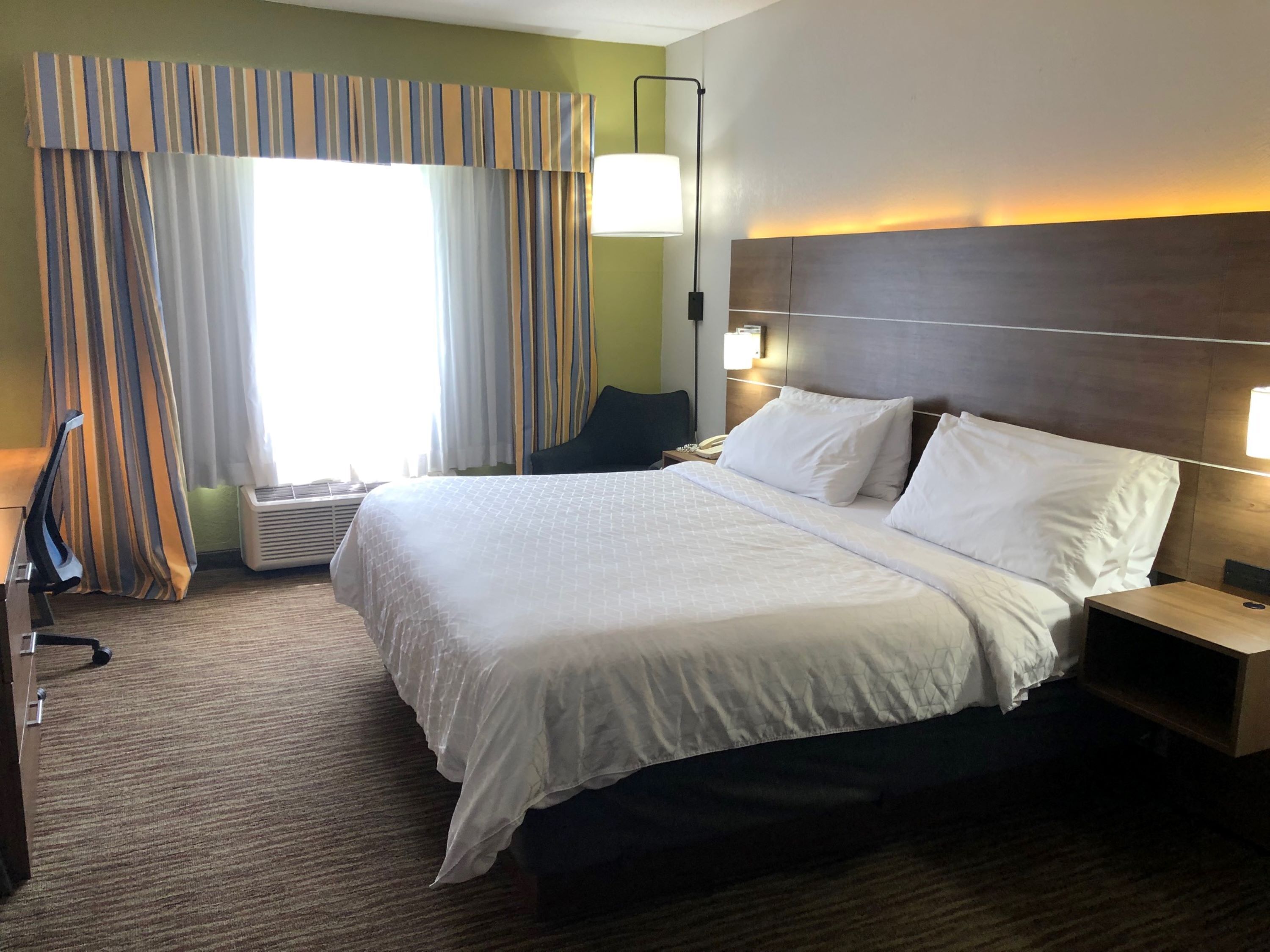 Une chambre d’hôtel avec un grand lit, une table de chevet, une lampe et une fenêtre avec des rideaux.