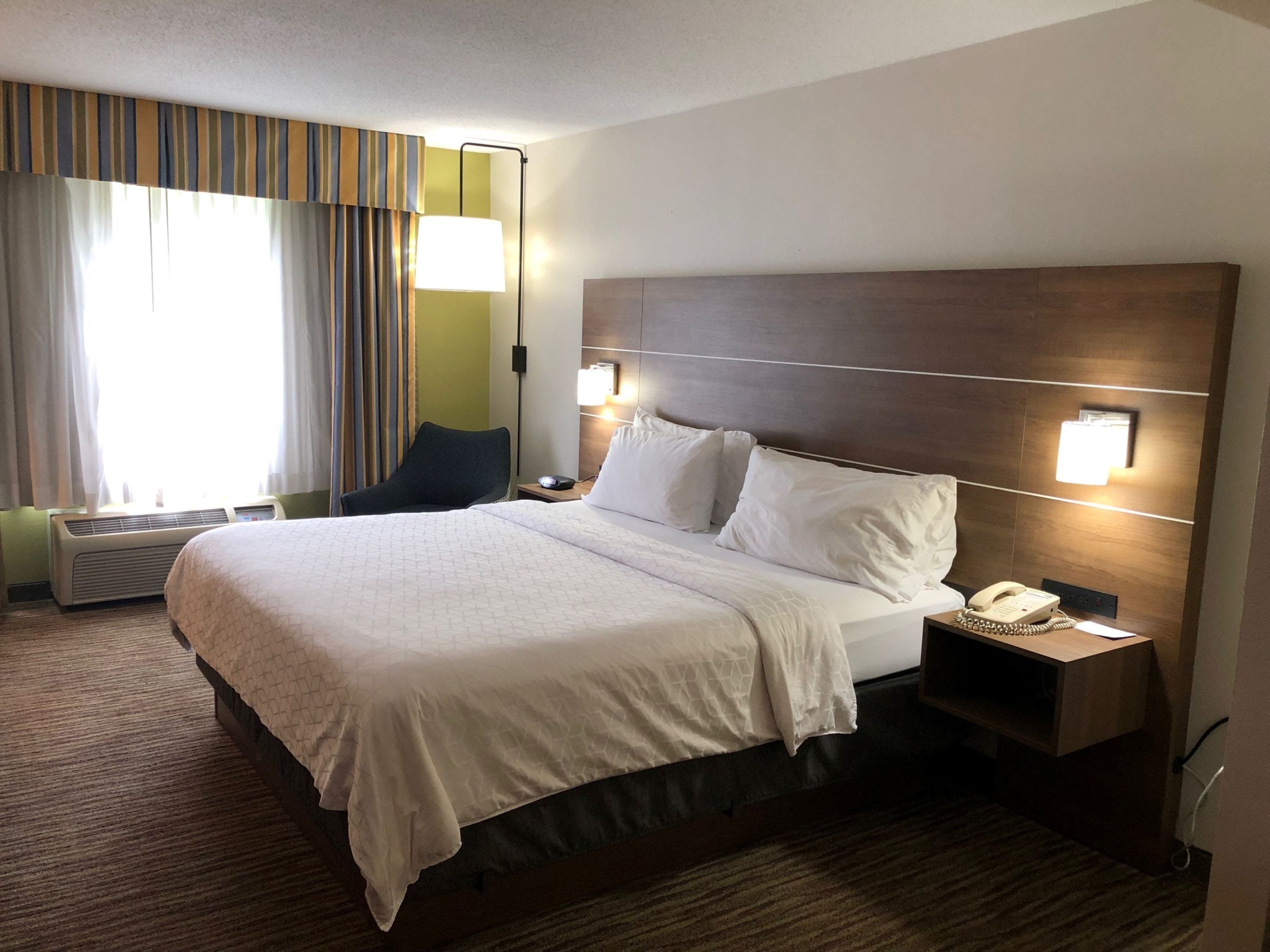 Habitación de hotel con una cama grande, una mesita de noche con lámpara, teléfono y ventana con cortinas.