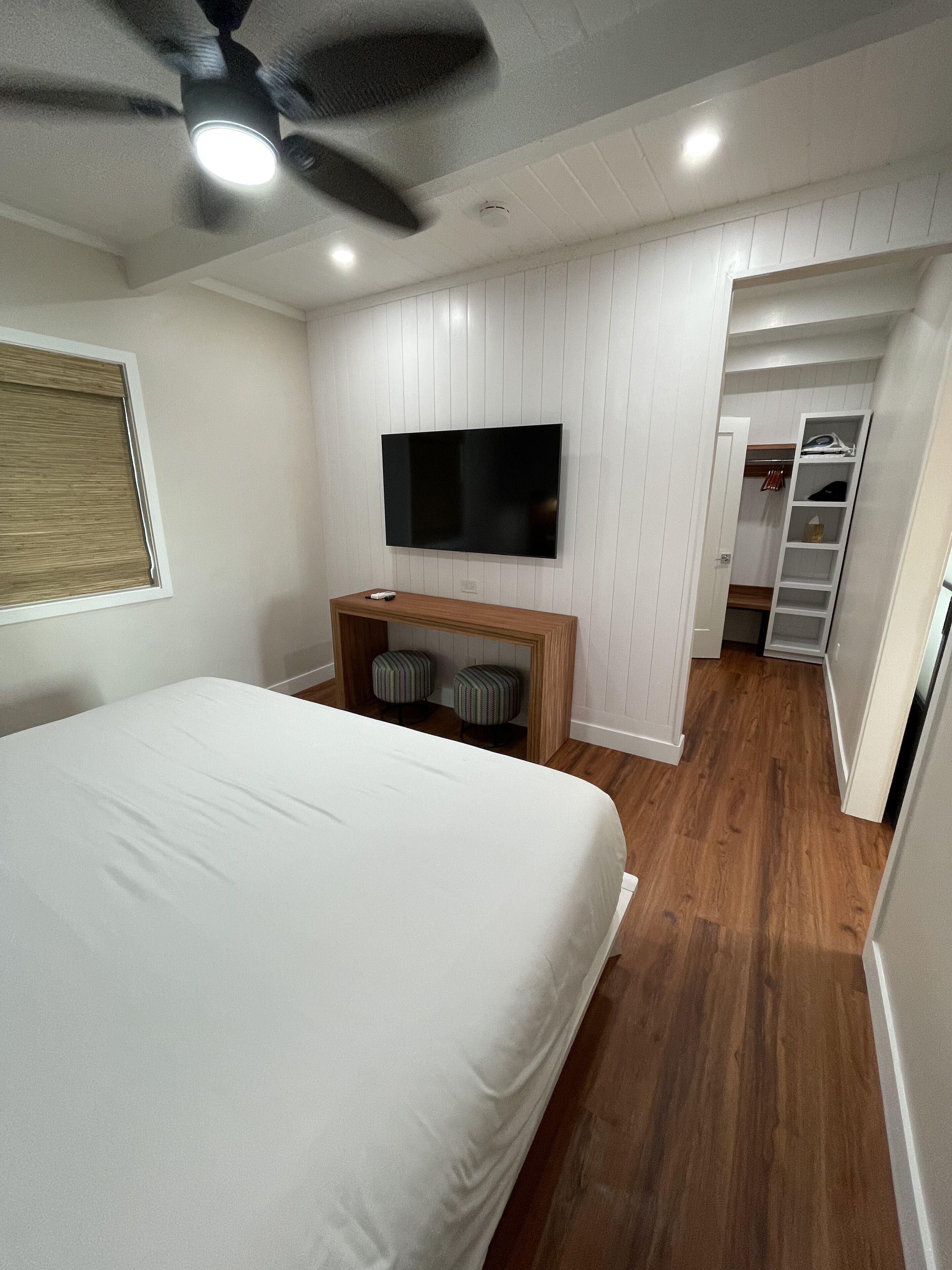 A bedroom with a bed, a TV mounted on the wall, a ceiling fan, and a wooden bench with two stools.