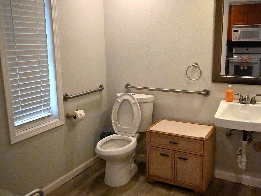 A bathroom with a toilet, wooden cabinet, sink, and a window with blinds.
