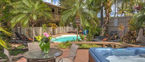 A pool area with lounge chairs, a hot tub, and a wooden deck surrounded by palm trees.
