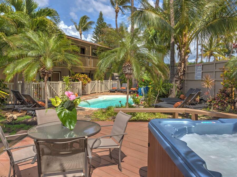 A pool area with lounge chairs, a hot tub, and a wooden deck surrounded by palm trees.