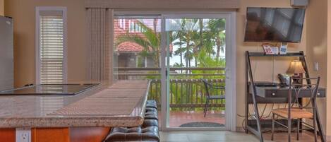 A kitchen with a large island, a dining area with a bench, a TV mounted on the wall, and a view of palm trees through the sliding glass doors.