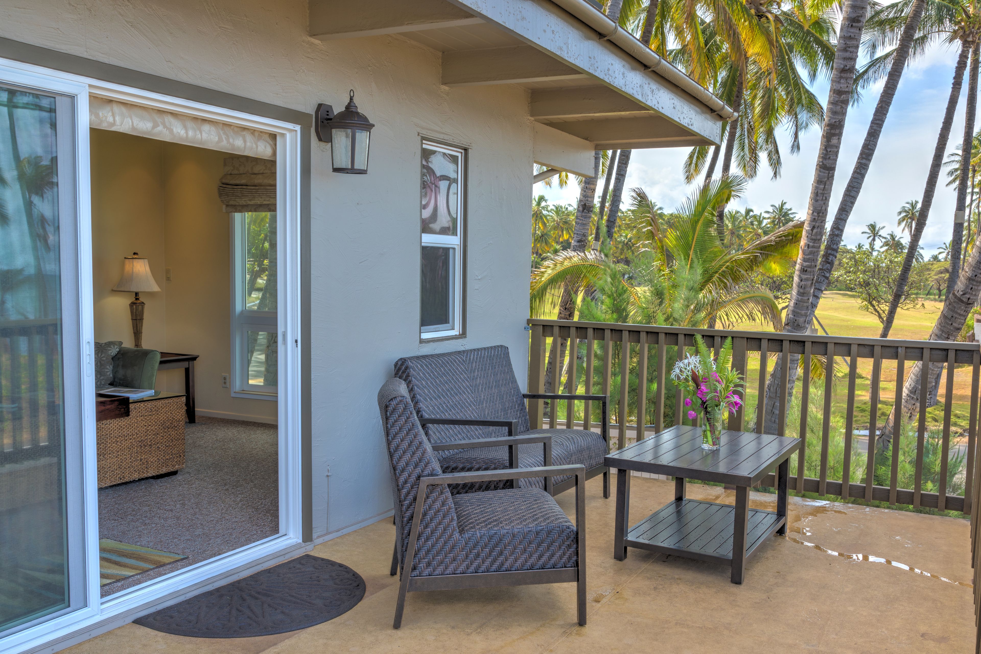 A balcony with a chair and table, a door leading to an interior room, and a view of palm trees and a golf course.