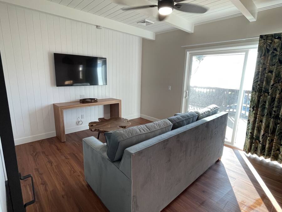 A modern living room with a grey sofa, wooden flooring, a ceiling fan, a flat-screen TV, and a large window with curtains.