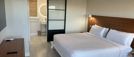 A hotel room with a bed, a desk, a ceiling fan, a bathroom with a mirror, and a view of the outdoors.