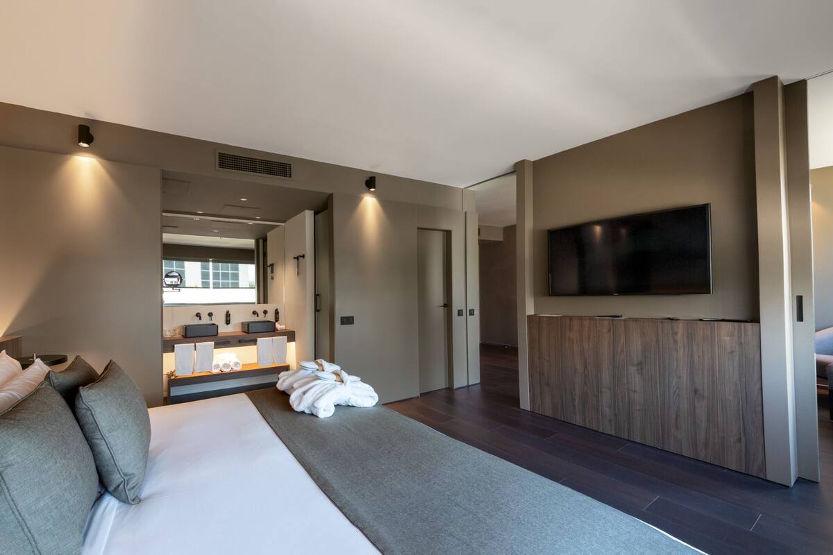 Quarto de hotel moderno com uma cama grande, uma televisão de tela plana fixada na parede, um banheiro com toucador e vista para o exterior.