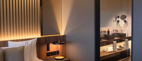 Una habitación de hotel moderna con cama, mesita de noche, lámpara y un baño visible a través de una pared de vidrio.