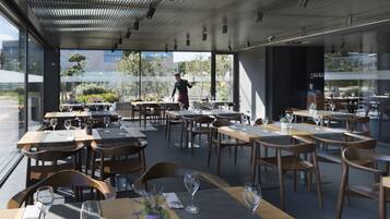 Moderni ravintola, jossa on puupöydät ja -tuolit, suuret ikkunat ja taustalla näkyy kävelevä henkilö.