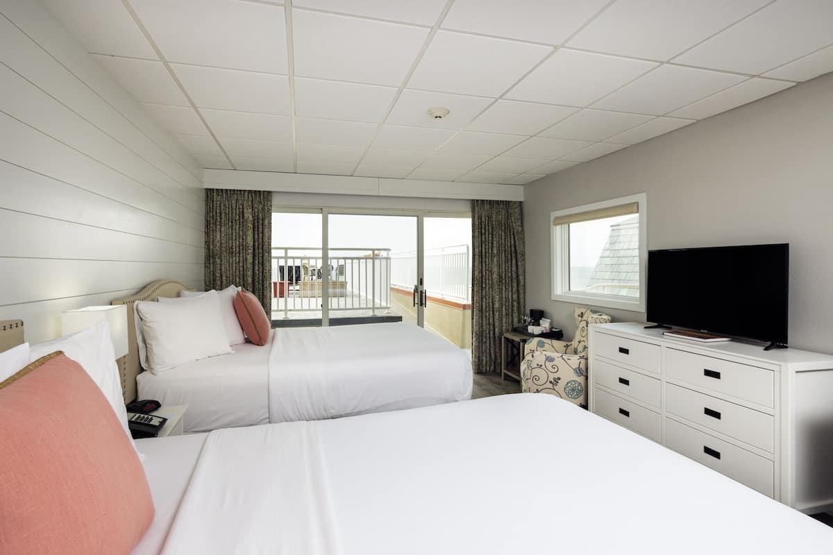 A hotel room with two beds, a TV, a desk, and a balcony with curtains.