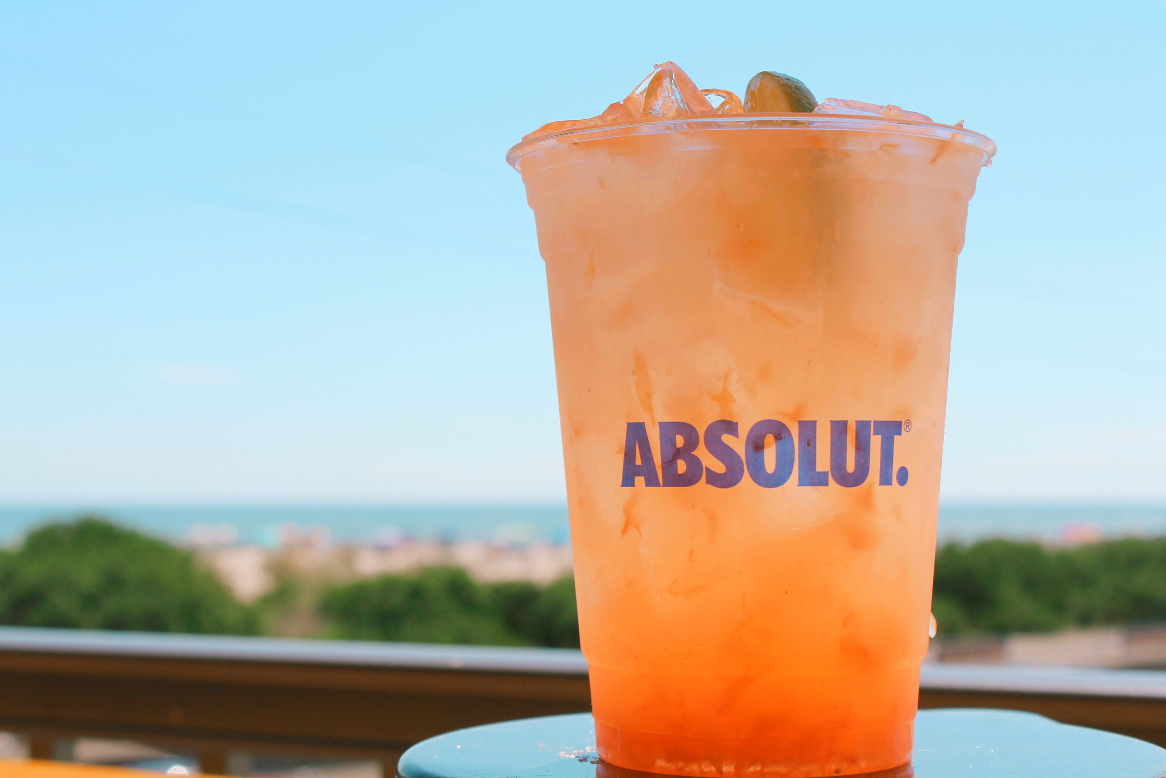 A plastic cup with the Absolut logo, filled with a red beverage, ice cubes, and a lime slice, placed on a table with a view of a cityscape in the background.