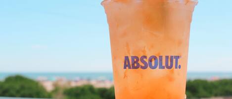 A plastic cup with the Absolut logo, filled with a red beverage, ice cubes, and a lime slice, placed on a table with a view of a cityscape in the background.