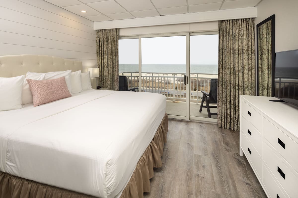 A hotel room with a large bed, a TV, a dresser, and a balcony with a view of the beach.