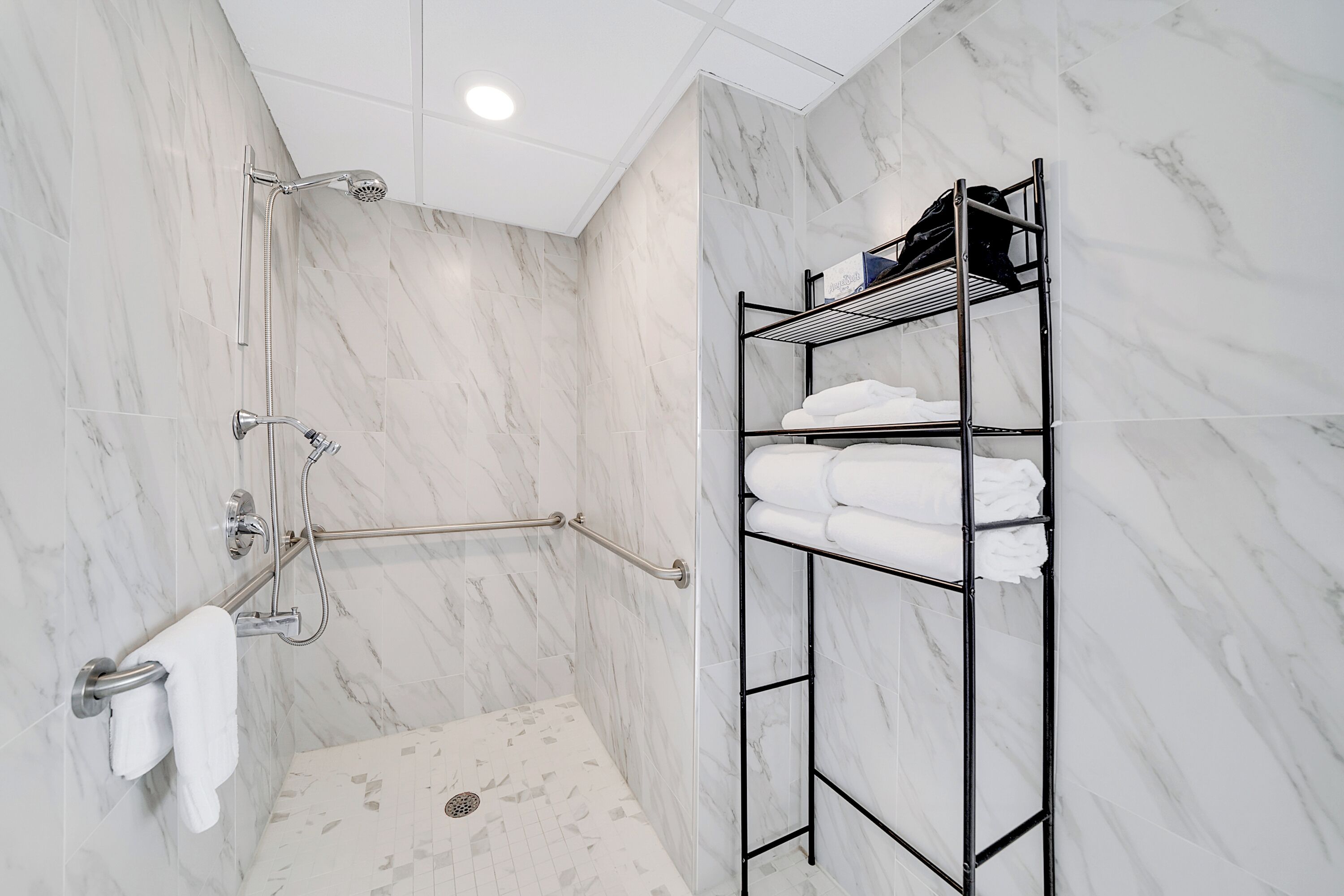 A modern bathroom with a walk-in shower, a grab bar, a towel rack, and a metal shelf with folded towels.