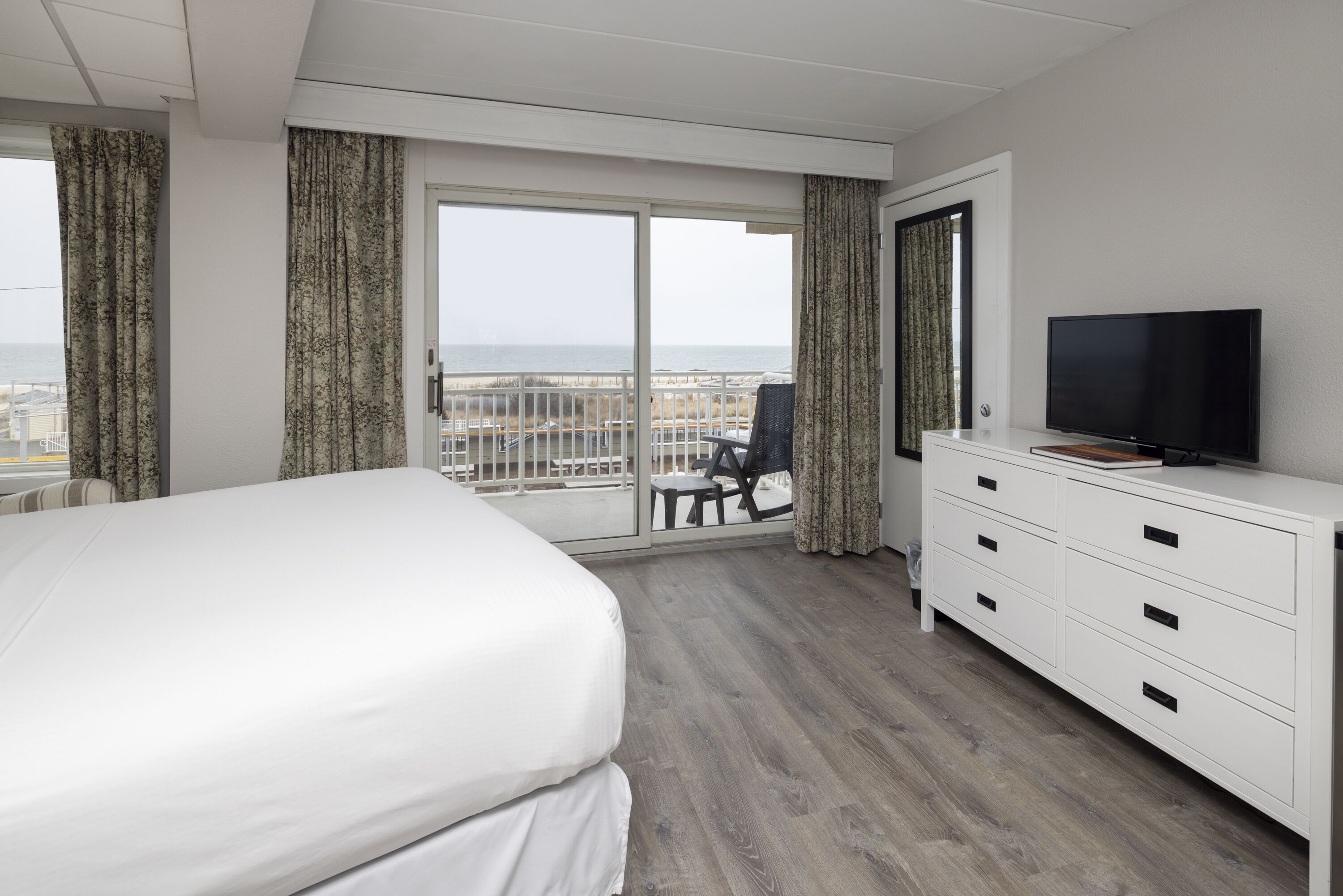 A hotel room with a large bed, a TV, a dresser, and a balcony with a view of the beach.