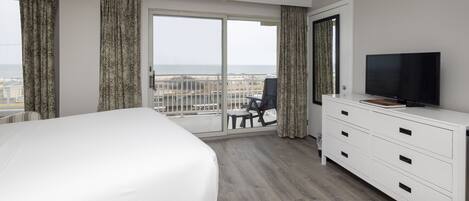 A hotel room with a large bed, a TV, a dresser, and a balcony with a view of the beach.