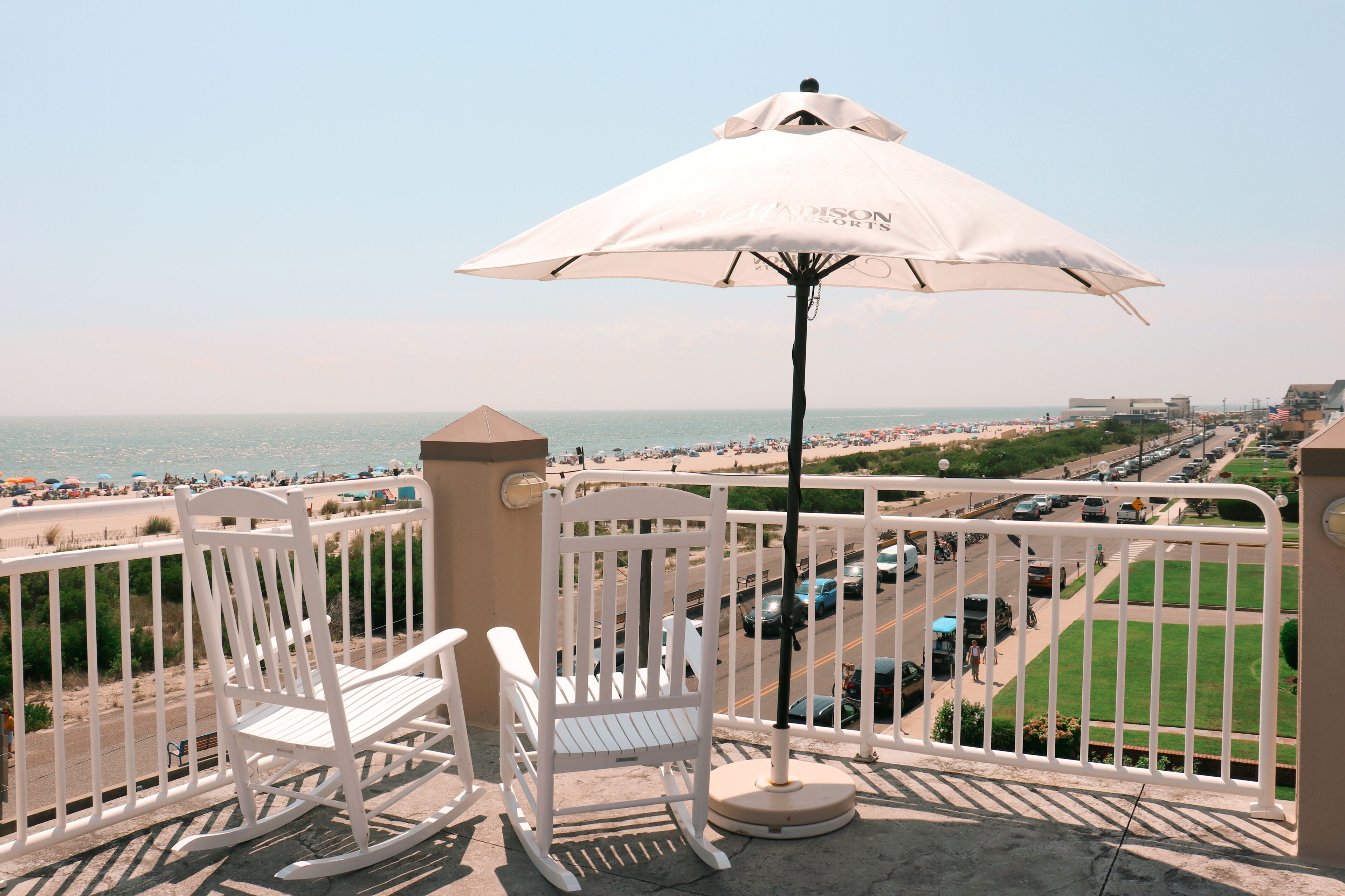 Un balcon avec un fauteuil à bascule blanc et un parasol, donnant sur une plage et une rue animée.