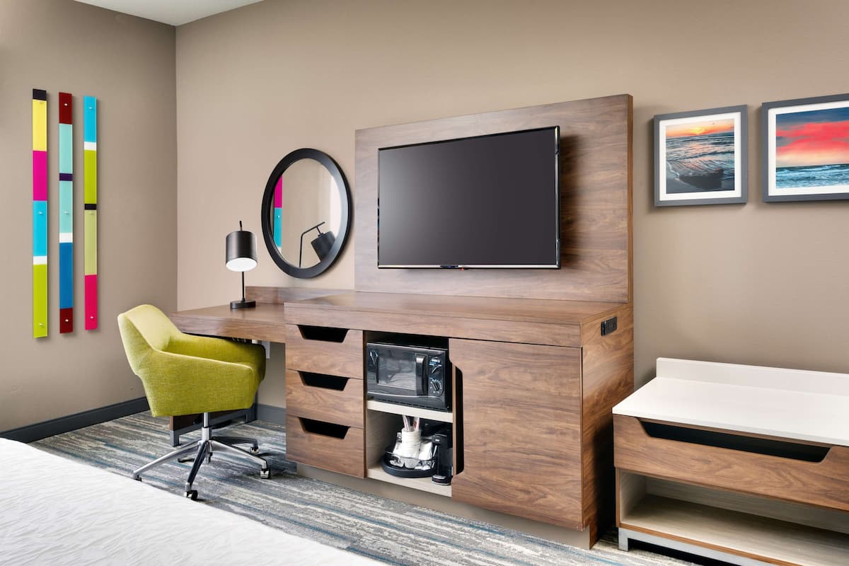 A hotel room with a wooden entertainment unit, a flat-screen TV, a green chair, a coffee machine, and a white desk.