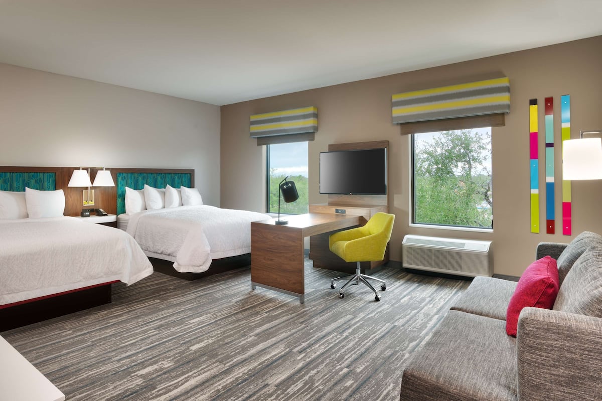 A hotel room with two beds, a desk, a chair, a TV, and large windows.