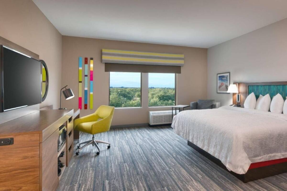 A hotel room with a bed, a desk, a chair, a television, a window with a view, and a wall-mounted air conditioner.