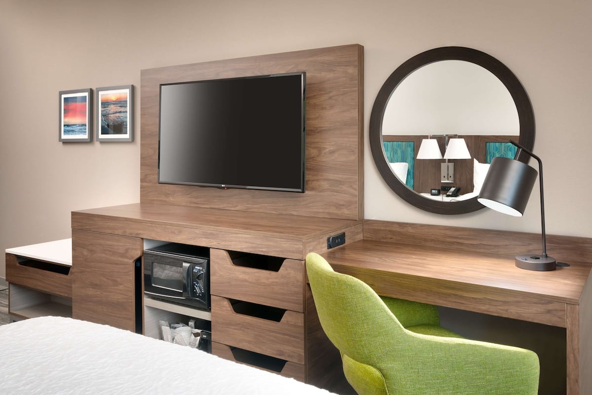 A hotel room with a flat-screen TV mounted on a wooden wall, a round mirror, a green chair, a desk with a lamp, and a bed with a white comforter.