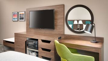 A hotel room with a flat-screen TV mounted on a wooden wall, a round mirror, a green chair, a desk with a lamp, and a bed with a white comforter.