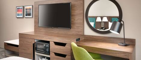 A hotel room with a flat-screen TV mounted on a wooden wall, a round mirror, a green chair, a desk with a lamp, and a bed with a white comforter.