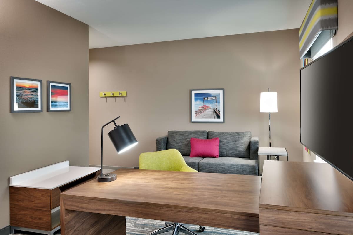 A modern hotel room with a wooden desk, a grey sofa, a flat-screen TV, and two framed pictures on the wall.