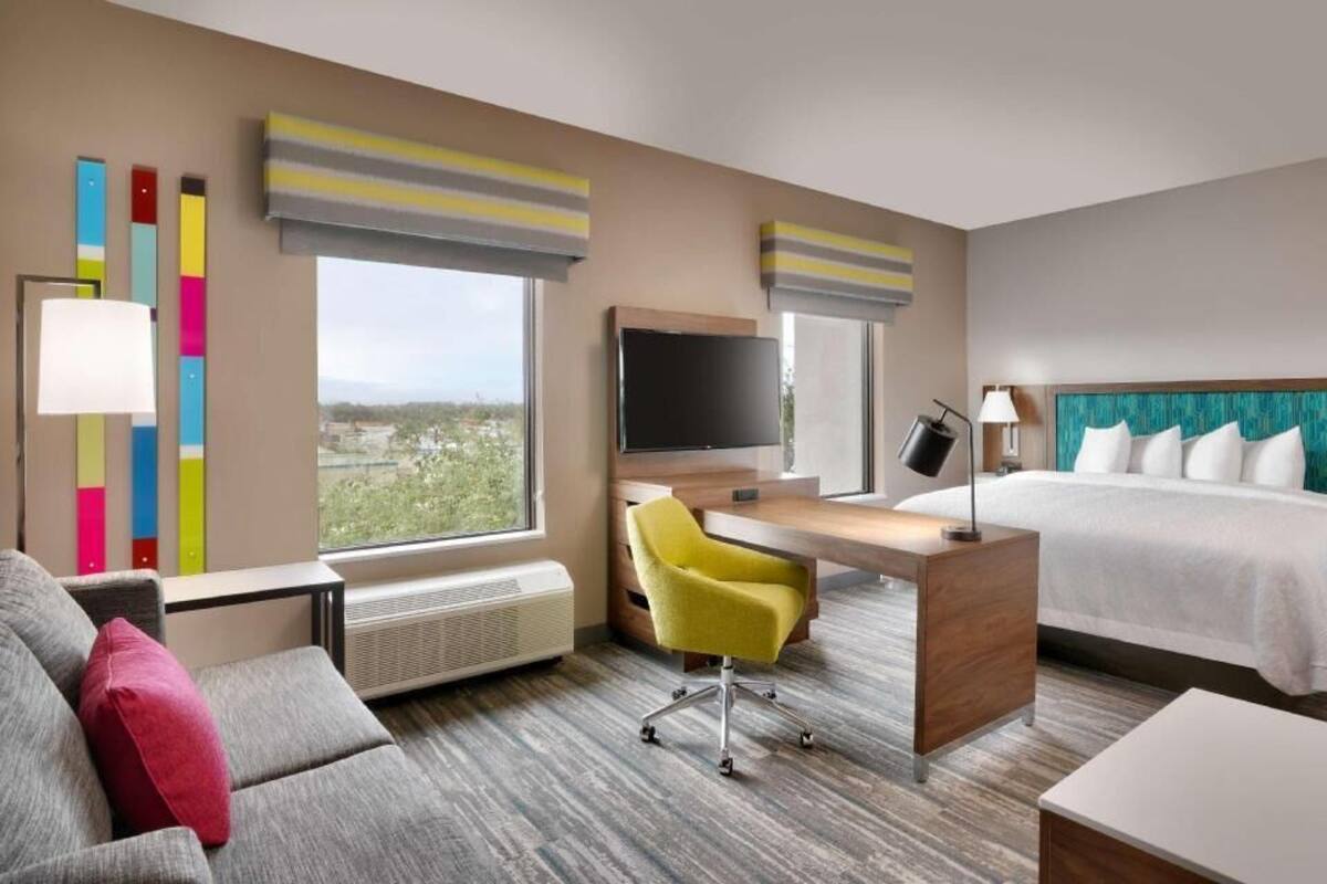 A hotel room with a bed, a desk, a chair, a TV, and a view of the outside.