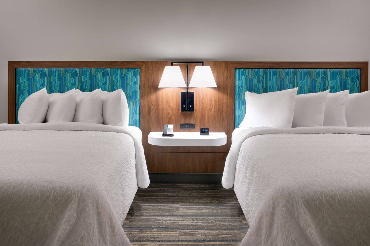 A hotel room with two beds, a bedside table with a phone and a digital clock, and a wall-mounted lamp.