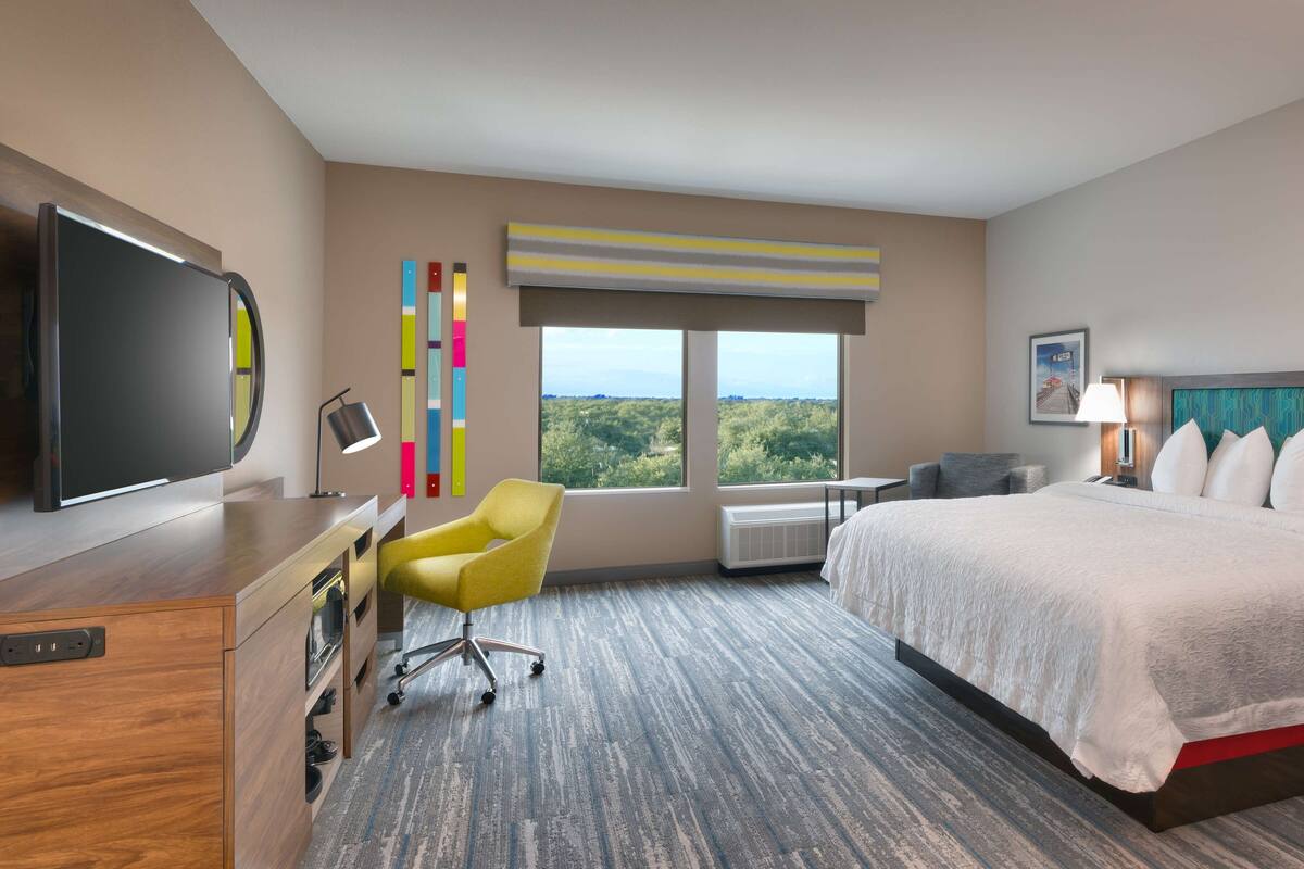 A hotel room with a bed, a desk, a chair, a TV, and a large window with a view of greenery.