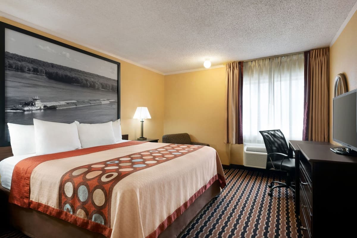 Habitación de hotel con cama, escritorio, silla, televisor, lámpara y un cuadro grande en la pared.