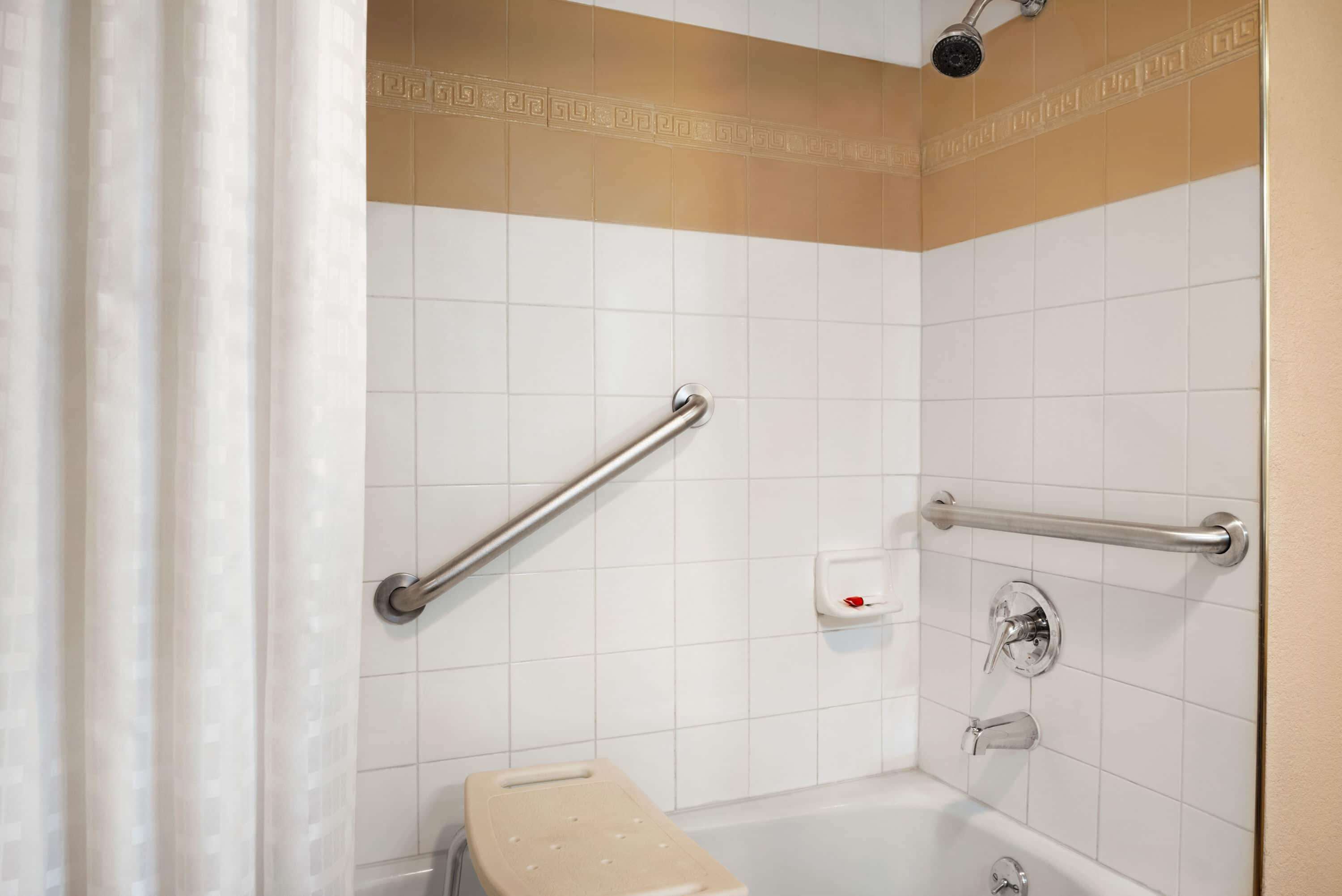 A bathroom with white tiled walls, a shower with a handheld showerhead, and two grab bars.