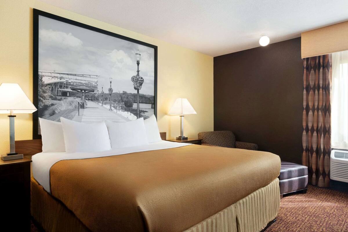 Una habitación de hotel con una cama grande, dos mesitas de noche, una silla, un cuadro grande enmarcado y un equipo de aire acondicionado.